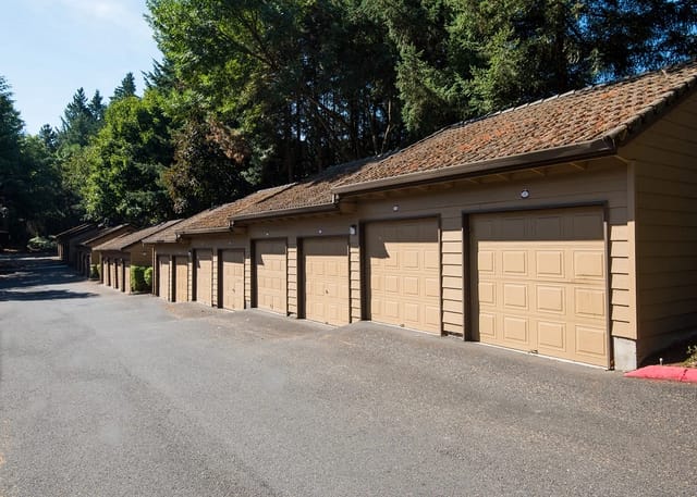 A long row of single-car garages with tan doors, set against a backdrop of lush green trees.