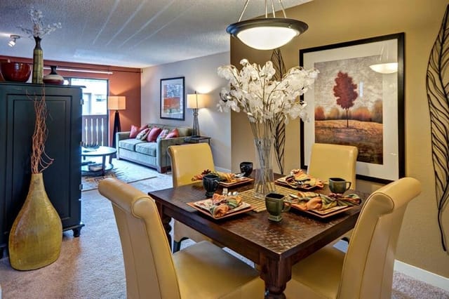 Dining room with a wooden table set for four, upholstered chairs, and a living room in the background.