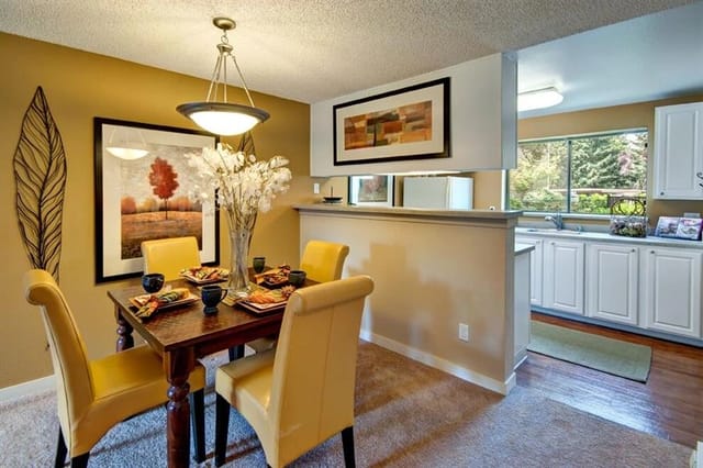Dining area with a table and chairs, adjacent to a kitchen with white cabinets and a window.