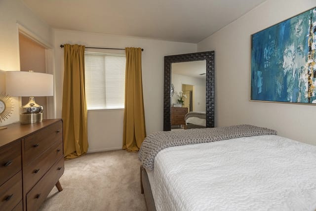 Cozy bedroom featuring a dresser with a lamp, yellow curtains, and a large mirror reflecting the bed.