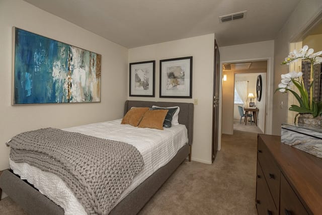 A cozy bedroom with a queen-sized bed, featuring plush bedding and decorative pillows. Artwork adorns the walls, and a doorway leads to an adjoining room.