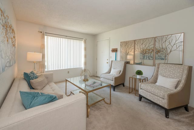 Bright living room in an apartment with a beige sofa, two patterned armchairs, glass coffee table, and wall art.