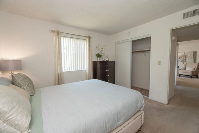 Bedroom in a neutral-toned apartment with a bed, nightstand, dresser, and sliding closet.