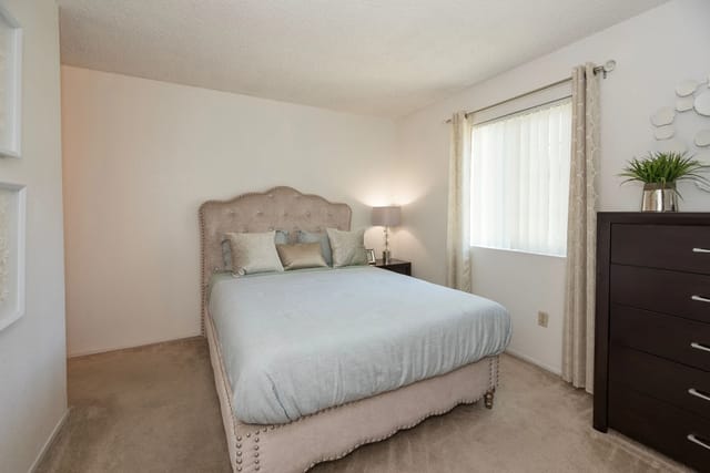 Bedroom with tufted upholstered bed, nightstand, dresser, and window blinds.