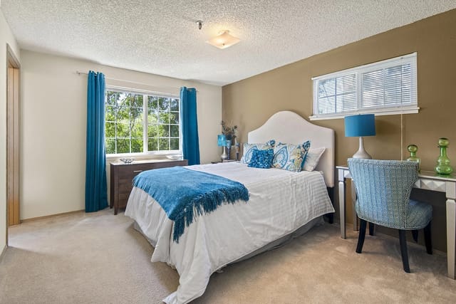 A bedroom with a white bed, blue accents, a desk, and a large window.