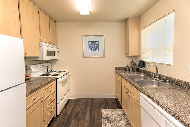 Apartment kitchen with light wood cabinets, white appliances, granite counters, and a double sink.