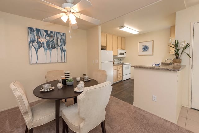 Dining area with a round table, four upholstered chairs, and a small kitchen in the background.