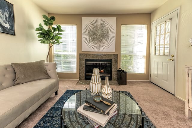 Living room in a modern apartment with a glass coffee table, sofa, fireplace, and large windows.
