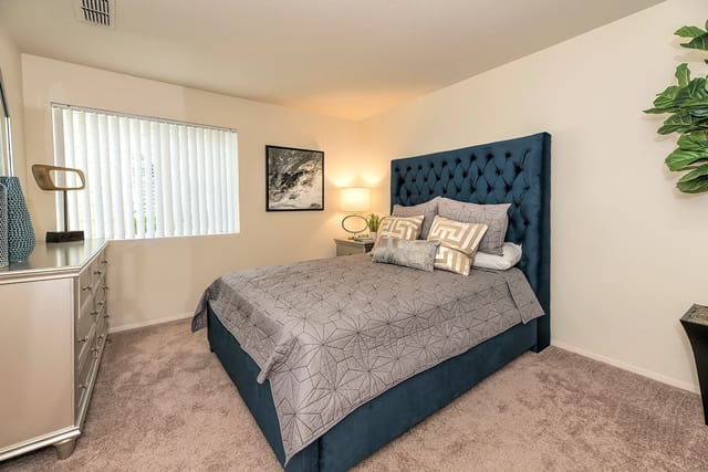 Bedroom with blue tufted headboard, grey geometric bedding, dresser, and lamp.
