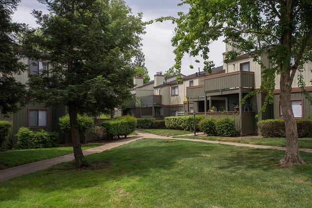 Exterior view of apartment buildings with green siding and balconies, surrounded by trees and grassy areas.