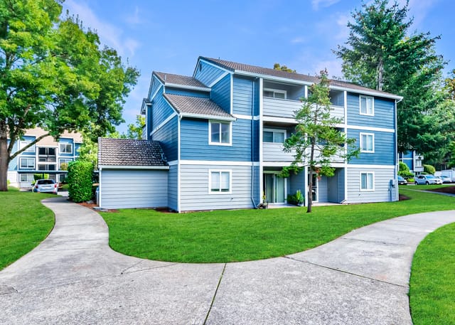 Exterior view of a blue apartment building with a paved walkway and lush green lawn.