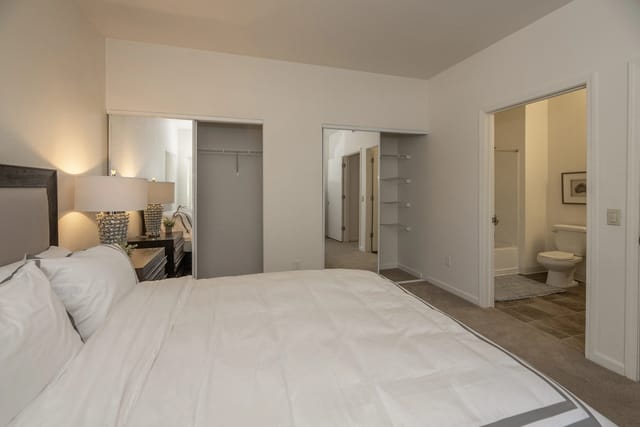 Bedroom with a bed, nightstand with lamps, and closet space. A bathroom is visible through an open doorway.