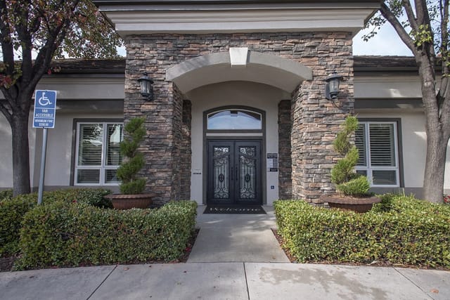 Building entrance with stone facade and double doors, flanked by bushes and trees.