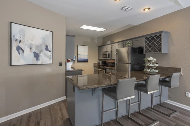 Modern kitchen island with granite countertop and bar stools, stainless steel appliances, and grey cabinets.