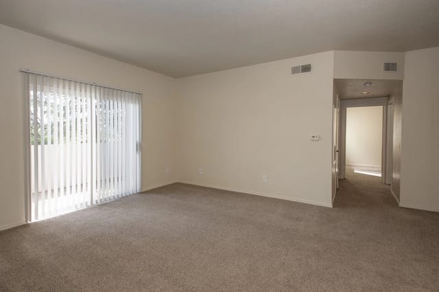 Spacious living room with carpet, vertical blinds on a sliding glass door, and an open doorway leading to another room.