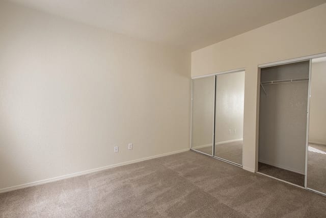 Interior view of an apartment bedroom with neutral walls, beige carpet, and sliding mirror closet doors.