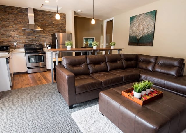 Modern living room with a sectional sofa, ottoman, and a stone accent wall behind the kitchen.