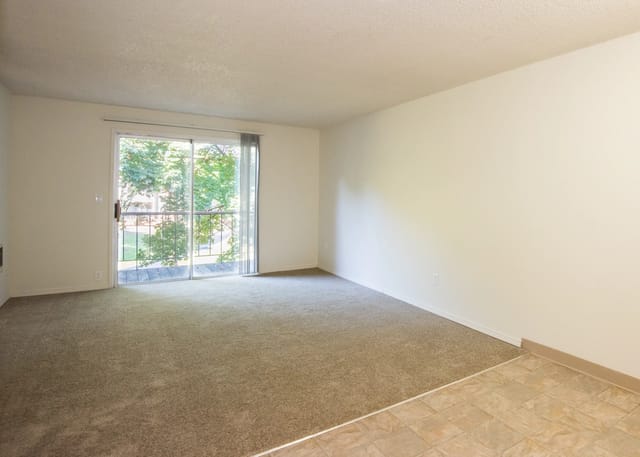Empty living room with carpet, sliding glass door to balcony, and tiled floor area.