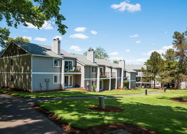 Exterior view of apartment buildings with green lawn and trees under a blue sky.