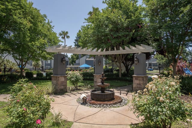 Fountain under a white pergola with stone pillars in a garden area.