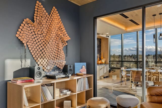 Interior of an apartment living area with wall art, a turntable, shelves, and a glass-walled kitchen.