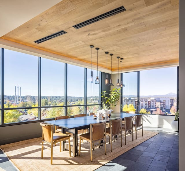 Communal dining area with a long table, tan chairs, pendant lights, and large windows showing city views.
