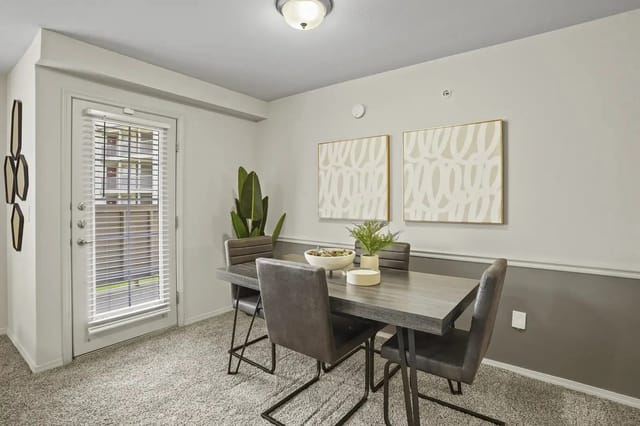 Dining area in a modern apartment with a gray table, chairs, plant, and wall art near a door with blinds.