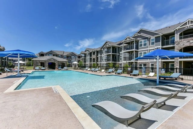 Outdoor resort-style pool with blue umbrellas, lounge chairs, and apartment buildings in the background.