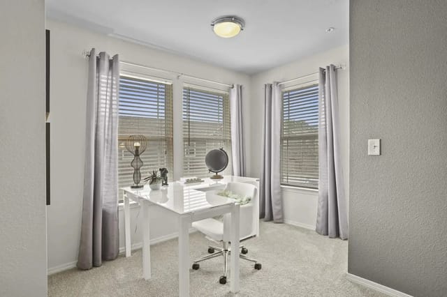 Bright apartment office with a white desk, chair, and three windows with blinds.