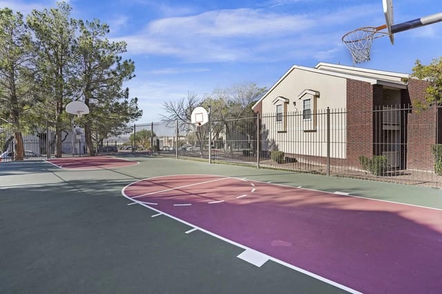 Outdoor basketball court with purple and green surface, fenced beside a brick building.