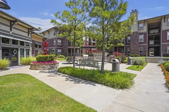 Outdoor community courtyard with trees, benches, and planters between apartment buildings.