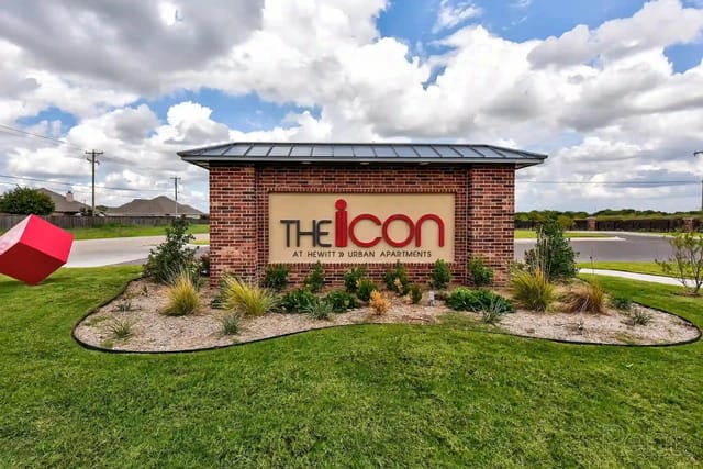Brick entrance sign for The Icon at Hewitt Urban Apartments with landscaping and blue sky.