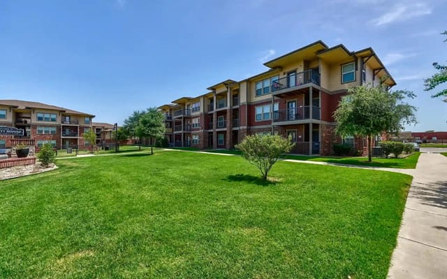 Exterior view of a multi-story apartment complex with balconies and a green lawn.