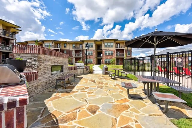 Outdoor community courtyard with stone patio, grills, and seating near apartment buildings.