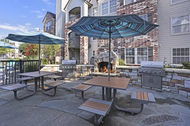 Outdoor communal patio with a stone fireplace, grills, umbrellas, and picnic tables.