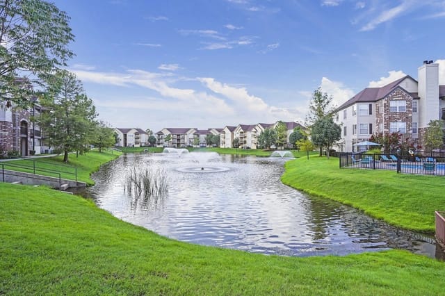 Exterior view of an apartment community with a central pond, fountains, and green lawns.