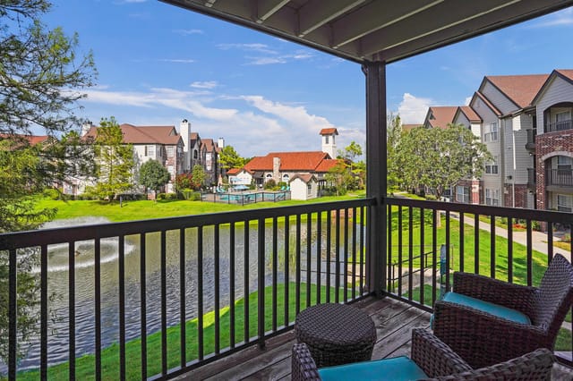View from balcony overlooking a pond with fountain, green lawn, and apartment buildings in the background.