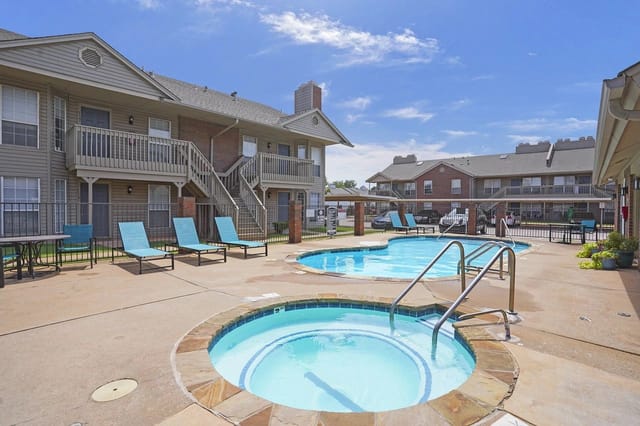 Outdoor pool and hot tub at an apartment community, with lounge chairs and surrounding residential buildings.