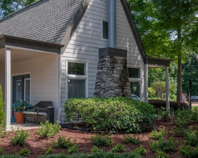 Exterior of a small apartment building with gray siding, stone chimney, and landscaped front yard.