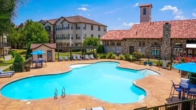 Outdoor pool with curved shape, lounge chairs, and blue umbrellas, backdrop of apartment buildings and a clubhouse.