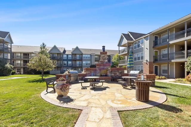 Community courtyard with a brick fireplace, seating, grills, and surrounding apartment buildings.