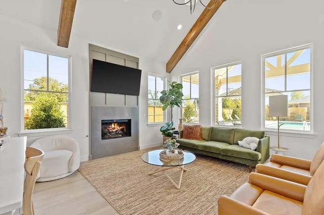 Bright living room with vaulted ceiling, exposed wooden beams, fireplace, wall-mounted TV, and large pool-view windows.