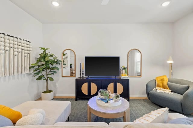 Bright living room with a TV on a dark cabinet, two arched mirrors, a large plant, and gray seating.