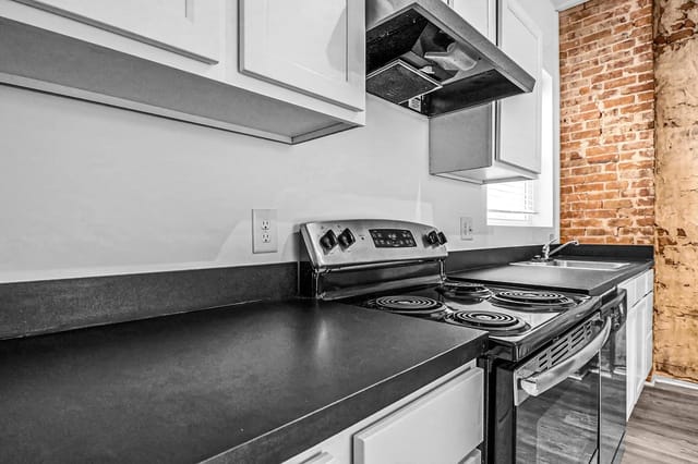 Close-up of a modern kitchen featuring a stainless steel range, black countertops, white cabinets, and exposed brick.
