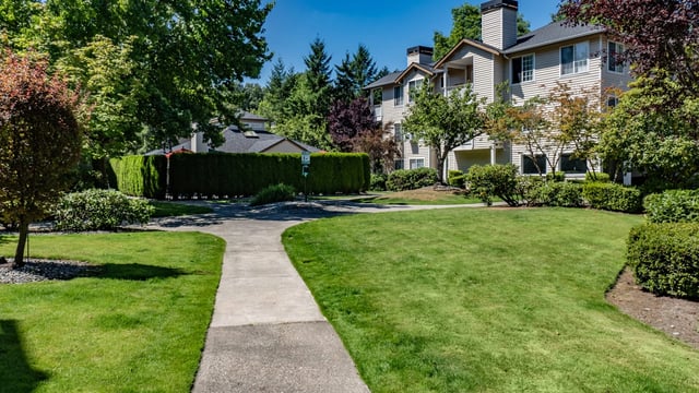 A paved walkway curves through a lush green lawn towards apartment buildings surrounded by trees and manicured hedges.