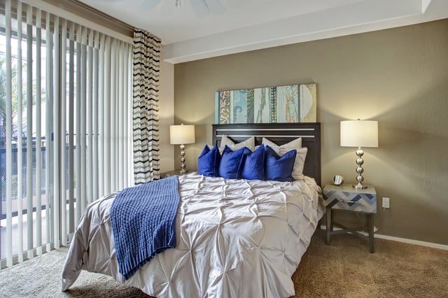 King-size bed with blue pillows and a blue throw in a bedroom with vertical blinds and a nightstand.
