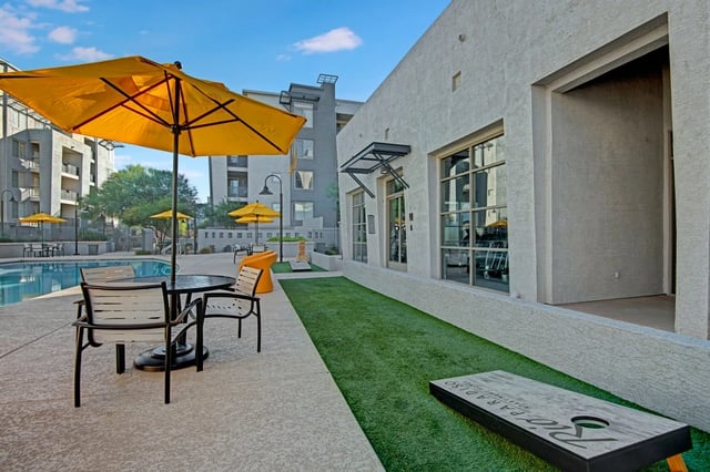 Outdoor seating area with a table and chairs next to a swimming pool with yellow umbrellas and a cornhole game.