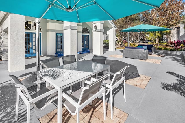 Outdoor seating area with white tables and chairs under a large teal umbrella.