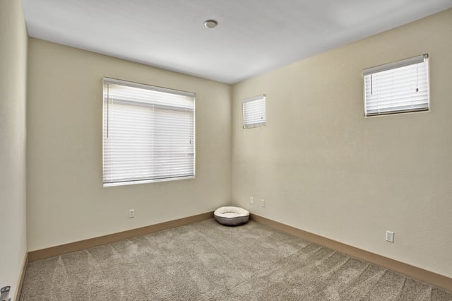 Empty apartment bedroom with neutral walls and carpet, and blinds on windows.