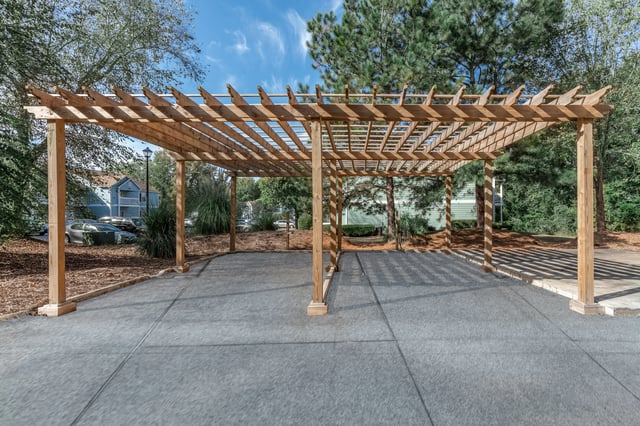Wooden pergola with concrete flooring and green trees in the background.